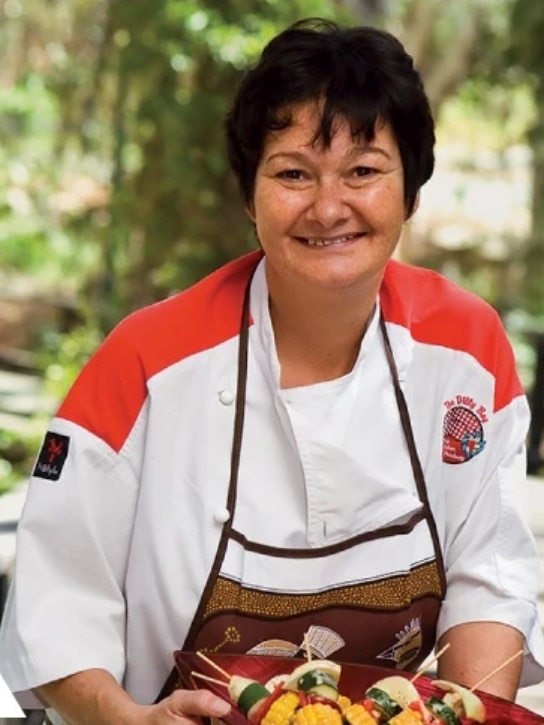 A woman in an apron holds a dish of vegetables.