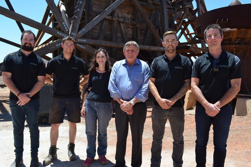 A group of six people standing in front of historic mine headframe.  