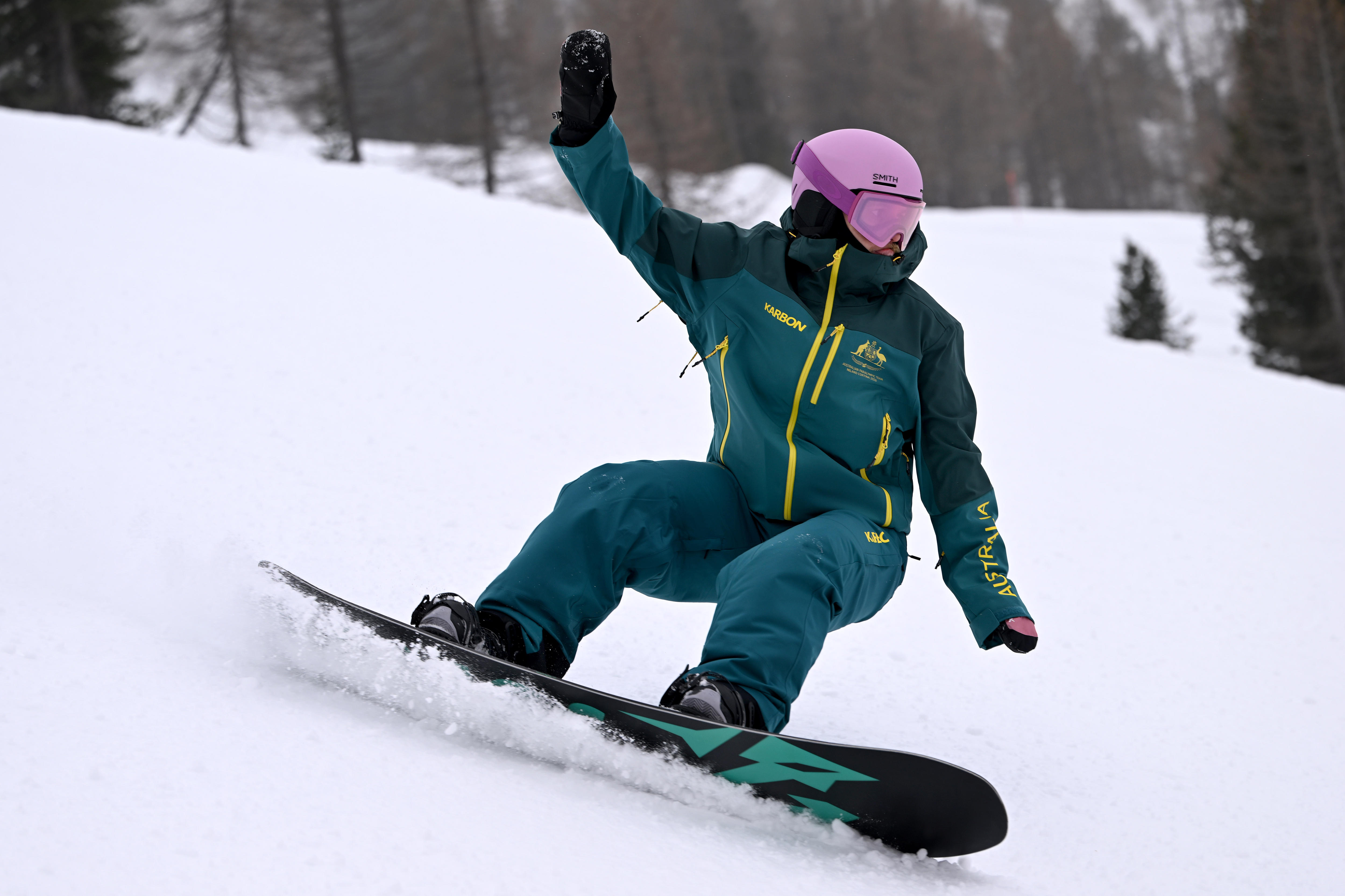 Um snowboarder vestindo o uniforme verde e dourado da Austrália e um capacete rosa está se inclinando para uma curva nas encostas.