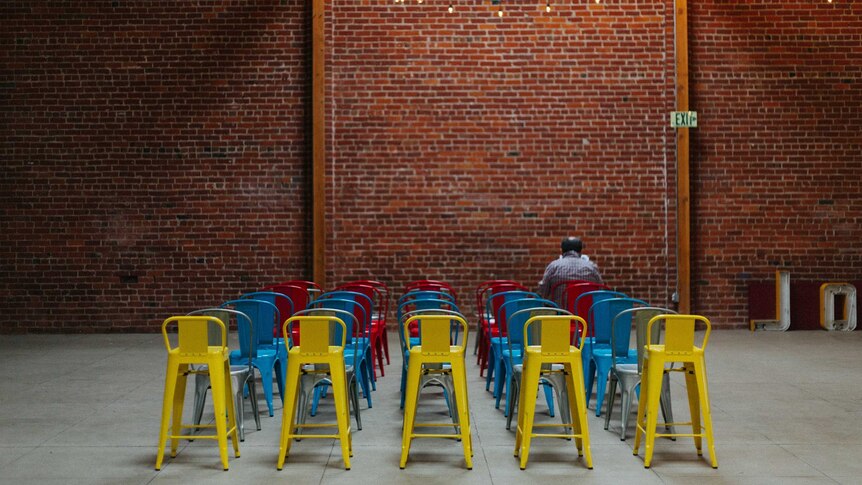 A man sits alone among a lot of empty chairs in primary colours.