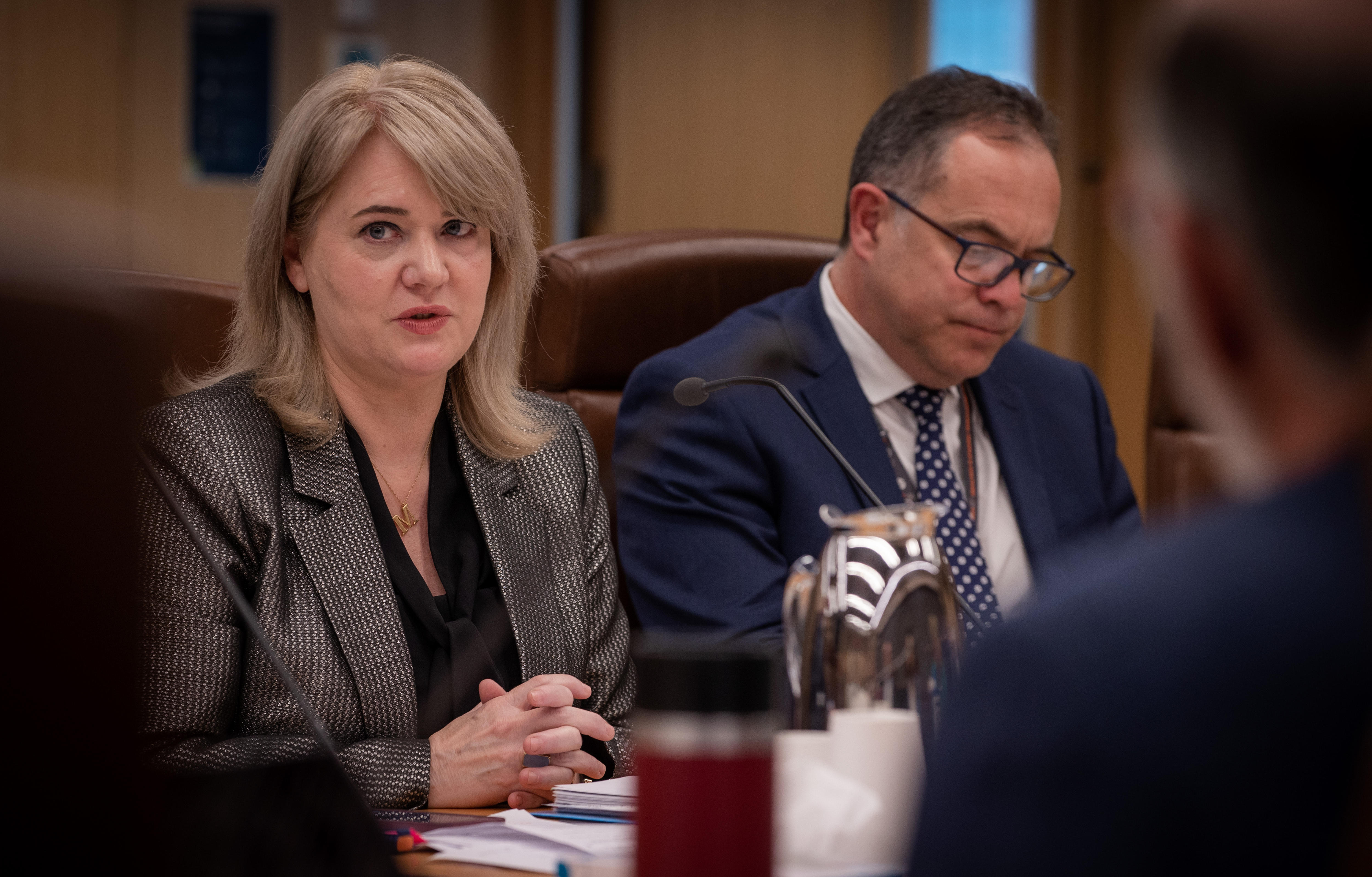A woman in a suit looks at the camera while a man in a suit, seated next to her, looks down at his papers 