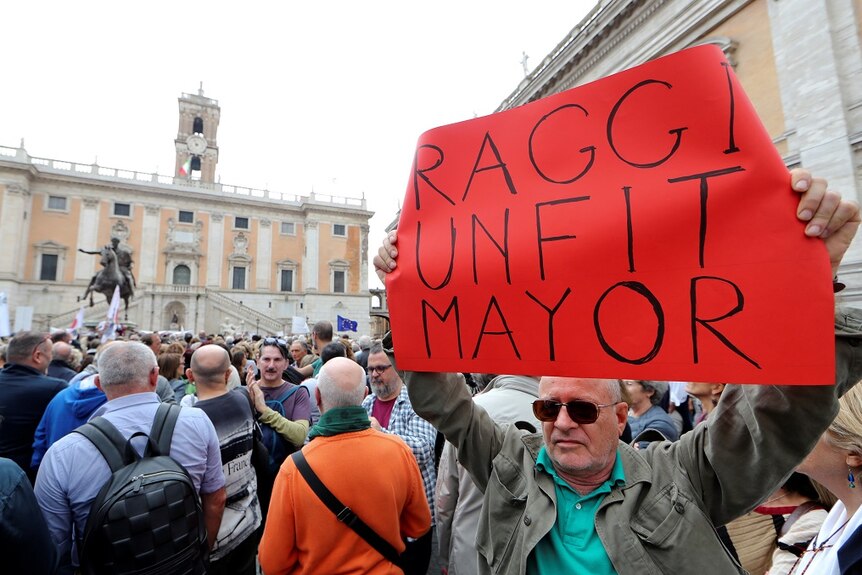A man standing in a packed crowd holds up a cardboard sign that says 'RAGGI UNFIT MAYOR'.