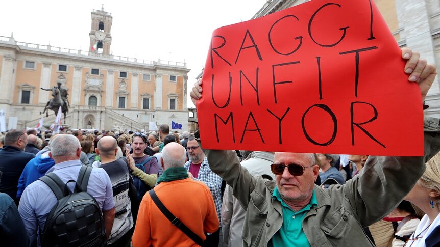 A man standing in a packed crowd holds up a cardboard sign that says 'RAGGI UNFIT MAYOR'.