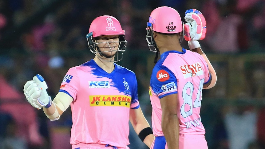 Steve Smith (left), wearing pink cricket equipment and helmet, grins and high fives a teammate