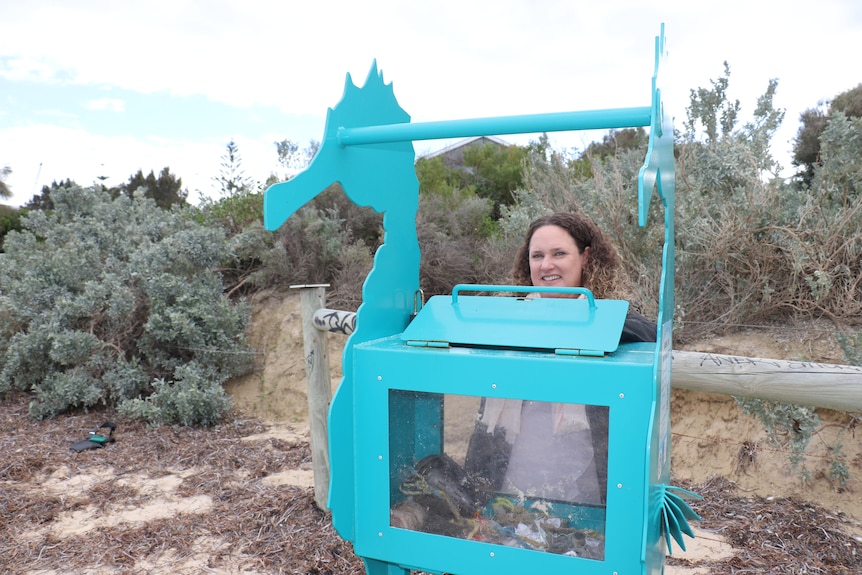 A light blue plastic bin with the design of a seahorse is seen on a beach. Behind, a middle-aged caucasian woman smiles.