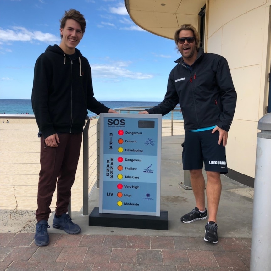 Two young men stand on a beach, beside a sign which indicates potential risks of sand banks, rips and UV radiation.