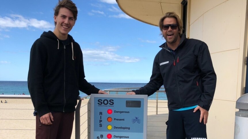 Two young men stand on a beach, beside a sign which indicates potential risks of sand banks, rips and UV radiation.