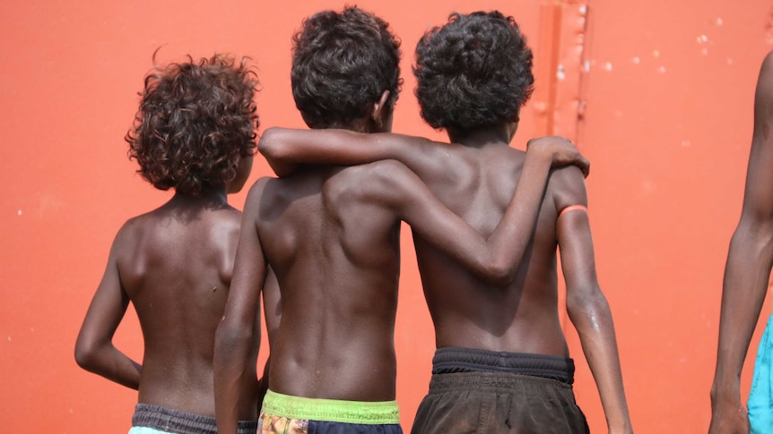 Three young shirtless Indigenous boys stand arm in arm with their backs to the camera in front of an orange wall.