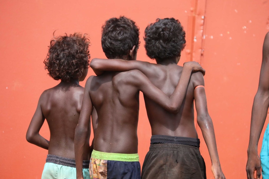 Three young shirtless Indigenous boys stand arm in arm with their backs to the camera in front of an orange wall.