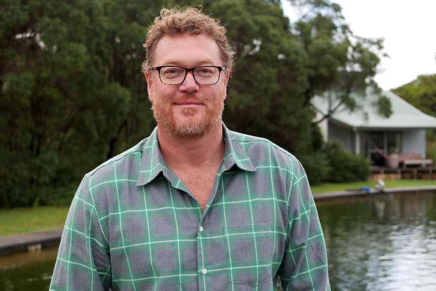 Head and shoulders shot of a red-haired man wearing glasses, with a lake and cottage in the background.