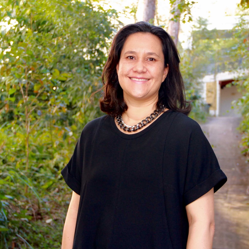 Woman with shoulder length hair with bulky neck chain wearing a black shirt stand in front of some trees.