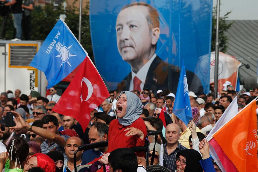 A woman in a wheelchair cries as she is lifted by protesters. There is a huge banner of Tayyip Erdogan behind her.