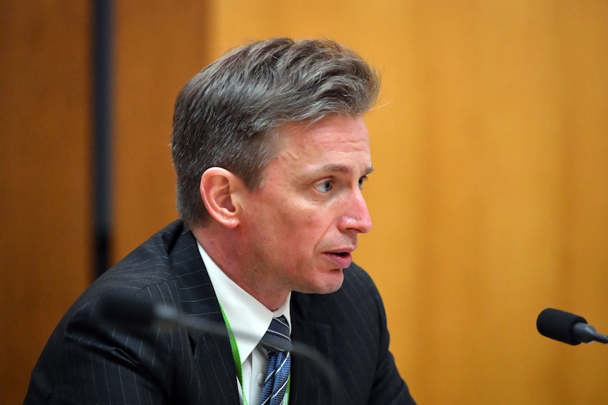 A middle-aged white man with grey hair wearing a suit speaks into a microphone in front of wood-panelled walls.