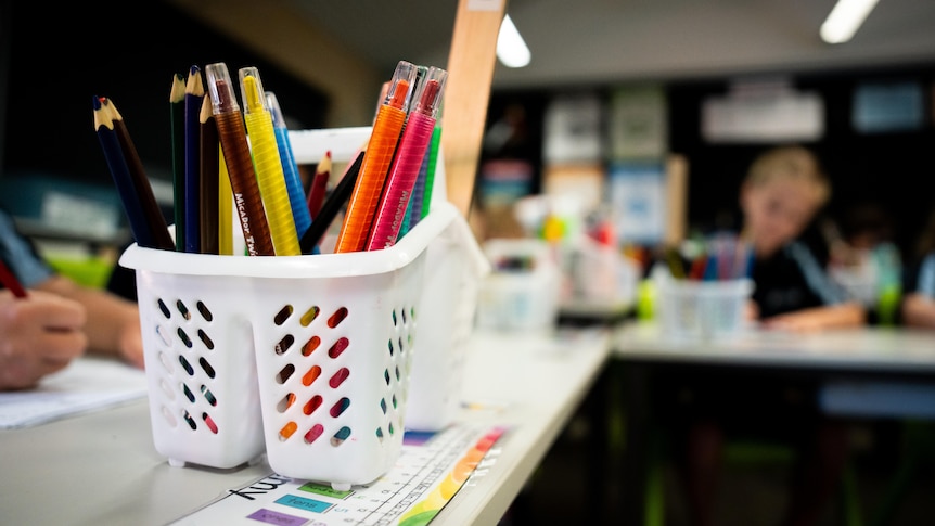 A close-up shot of pencils in a white container on a school desk, with blurred students in the background.