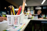 A close-up shot of pencils in a white container on a school desk, with blurred students in the background.
