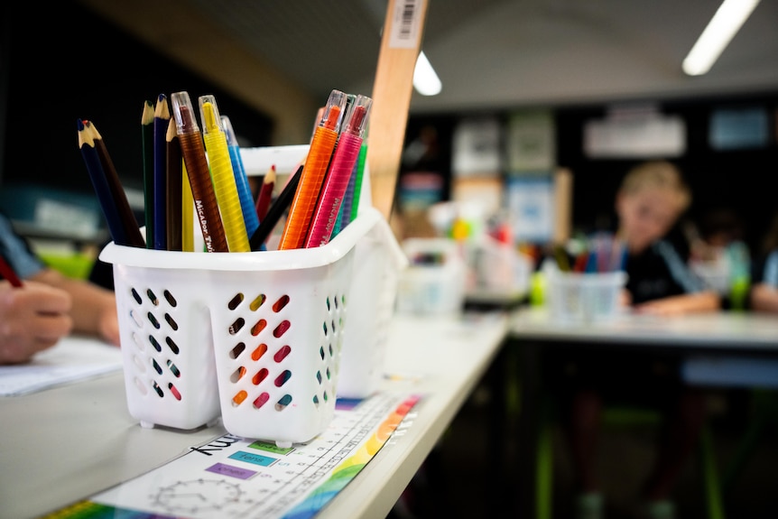 A close-up shot of pencils in a white container on a school desk, with blurred students in the background.