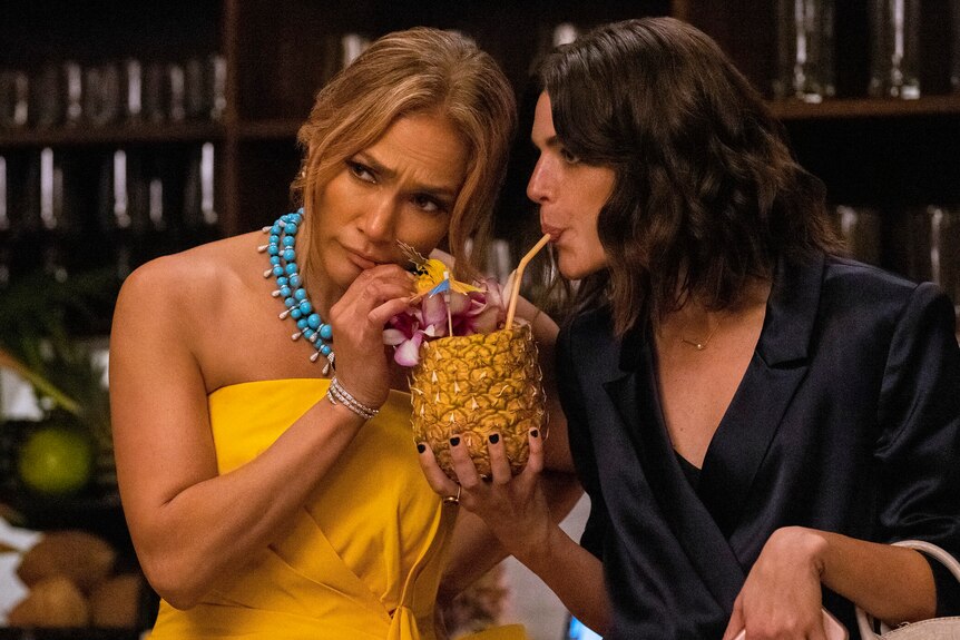 A Latina woman with honey hair wears a yellow dress and drinks from a pineapple with a brunette Latina in black silk.