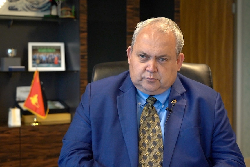 A man with grey hair wearing a blue suit with gold and black tie sits in an office chair.