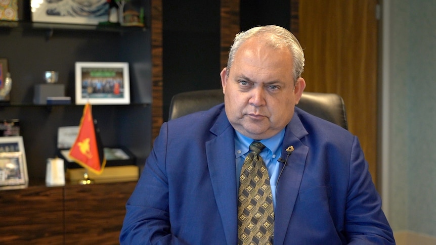 A man with grey hair wearing a blue suit with gold and black tie sits in an office chair.