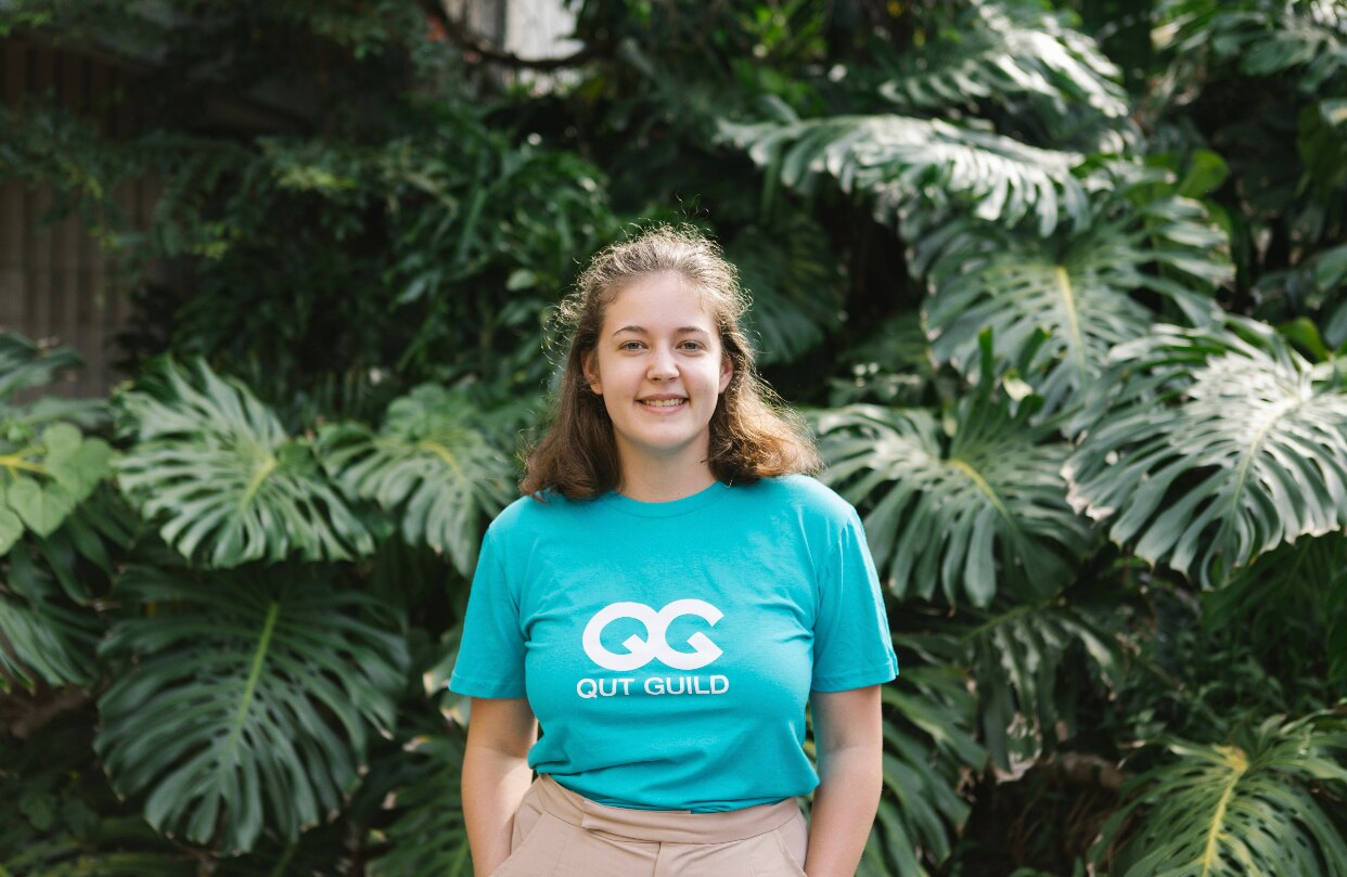 A young woman standing in front of a grove wearing a QUT Guild t-shirt.