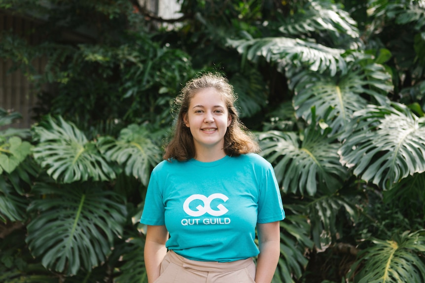 A young woman standing in front of shrubbery with a QUT Guild shirt on.