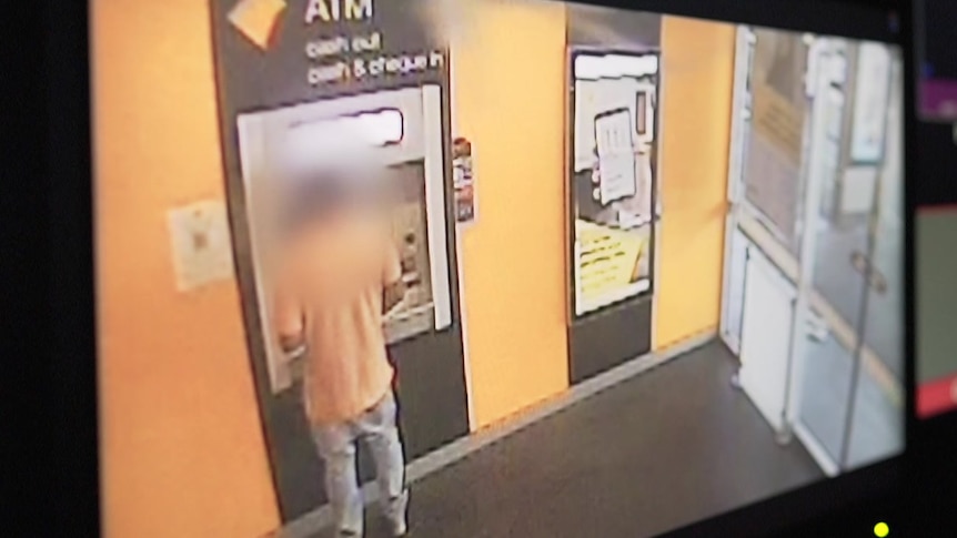 A man stands at a Commonwealth Bank ATM. He wears an orange shirt and jeans. His face is blurred.