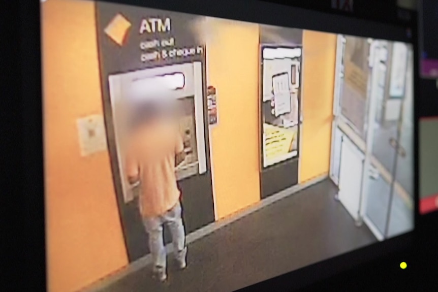 A man stands at a Commonwealth Bank ATM. He wears an orange shirt and jeans. His face is blurred.