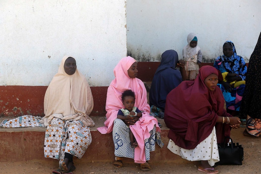 Women sit and wait, some holding babies, outside a building.