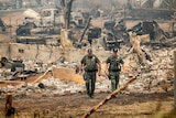 Two men in uniform walk in front of a house destroyed by fire. 