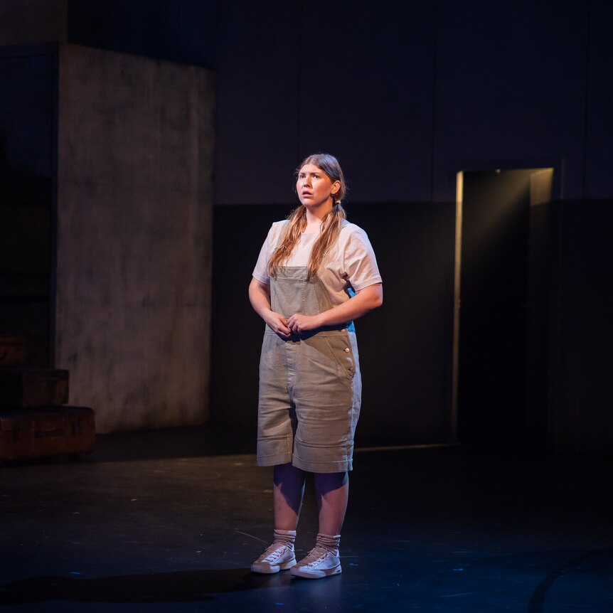 Woman on empty dark stage. Hair in low pigtails, beige t-shirt and overalls, white socks and converse. Hands touch infront 