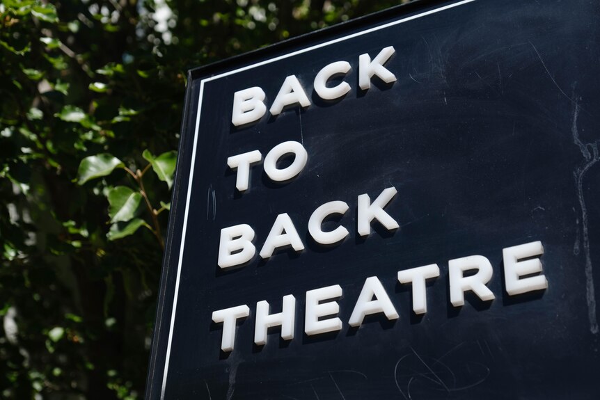 A back sign with white writing says Back to Back Theatre and is outside on a sunny day with a tree behind it