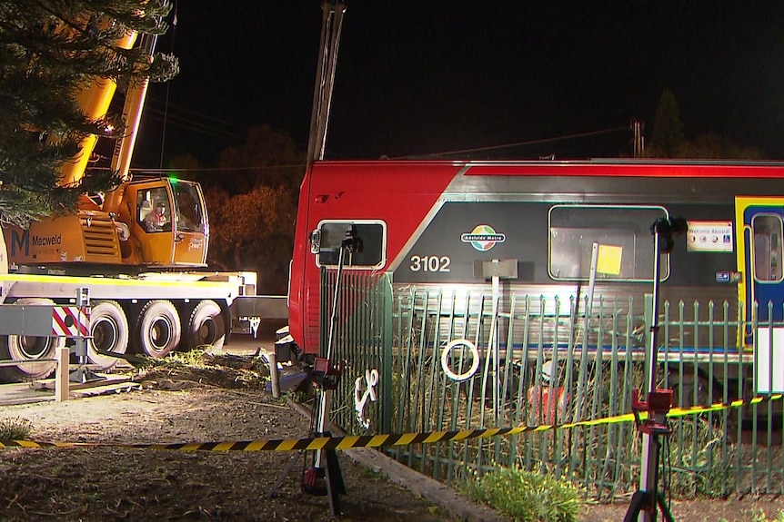 A yellow crane is parked next to a red train at night, the area is taped off