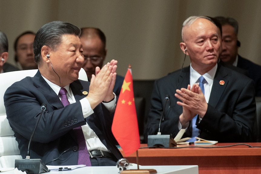 Xi Jinping claps as he wears an earpiece while sitting at a table on which a small Chinese flag sits.