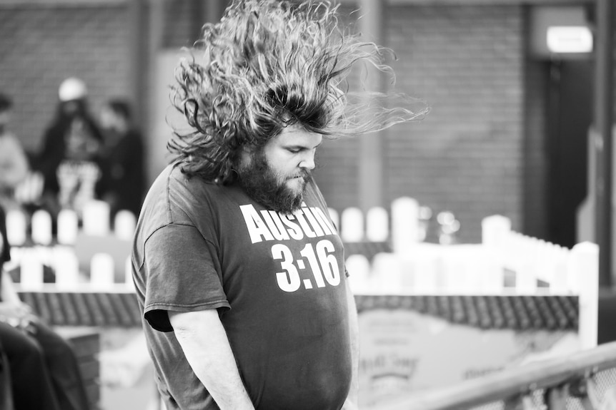 A man in his 30s with a beard and long hair flying has his eyes closed while he stands outdoors listening to music 