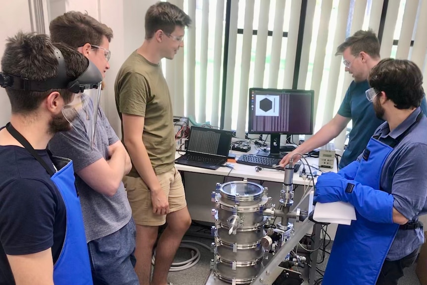 A group of students in a lab are gathered around a device testing their satellite