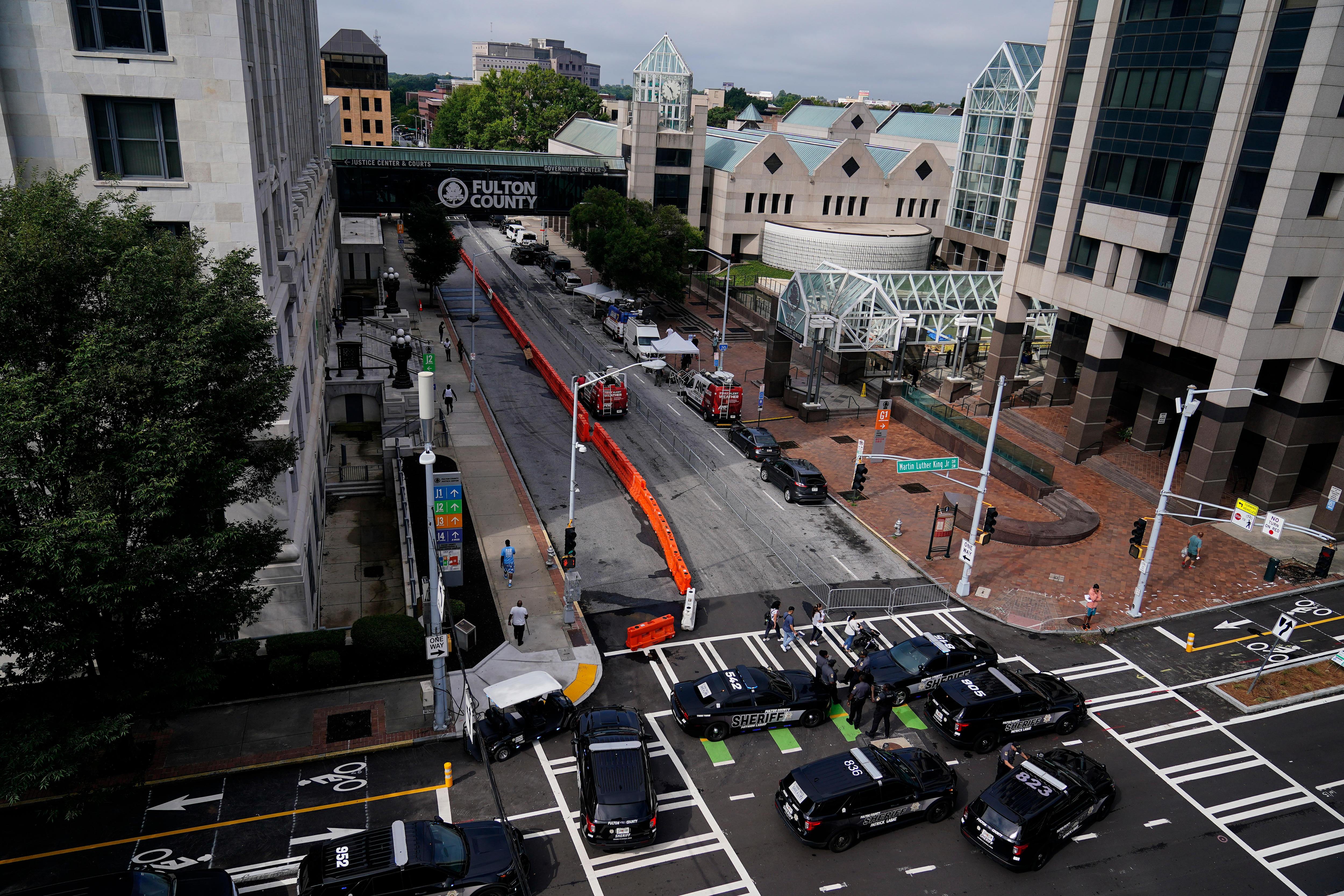 Seven police cars are parked in the middle of a road. Steel fencing and orange barricades line the sides of the street. 