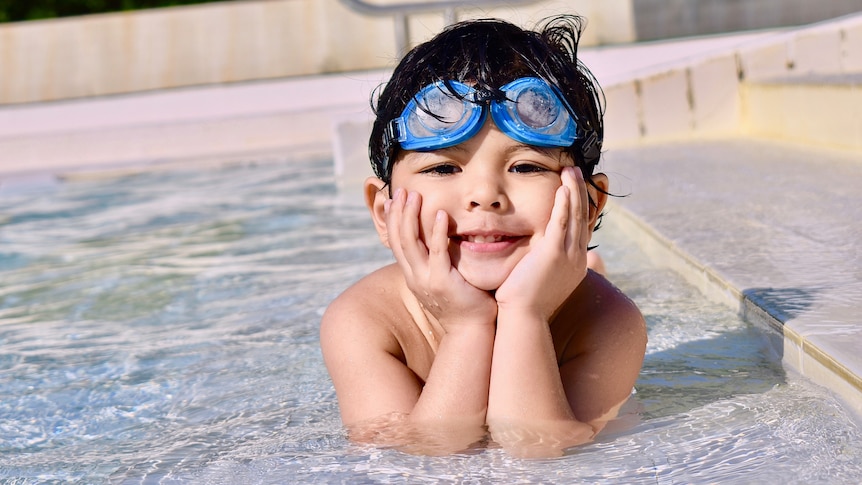 A small boy lies in the water of a shallow pool with his face in his hands for a story on keeping kids safe in the heat