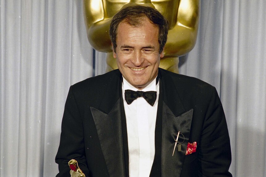 Bernardo Bertolucci stands in front of an Oscars statue while holding his Oscar.