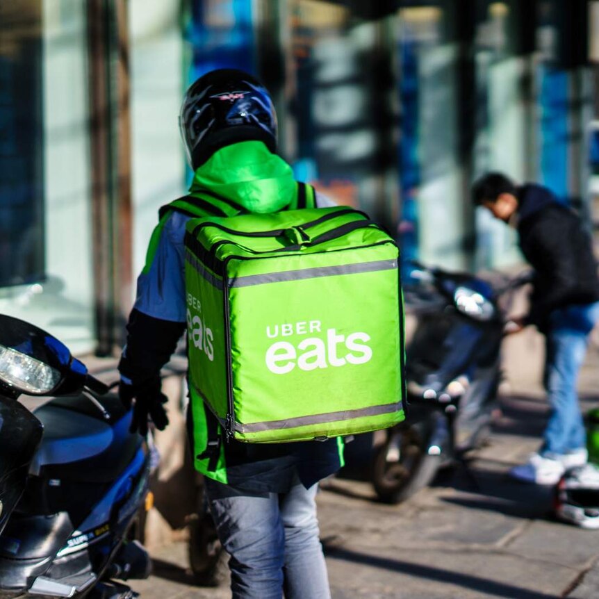 A courier wears a helmet and stands near a motorcycle, carrying a green Uber Eats food bag, with another courier in background