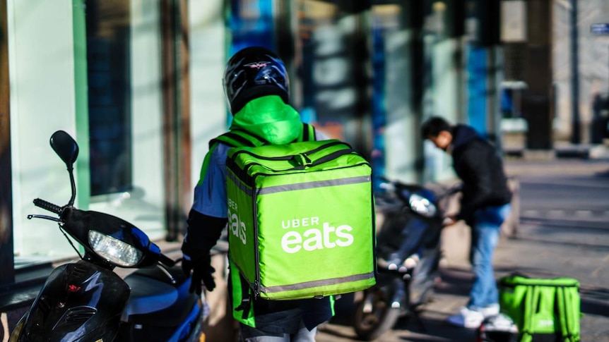 A courier wears a helmet and stands near a motorcycle, carrying a green Uber Eats food bag, with another courier in background