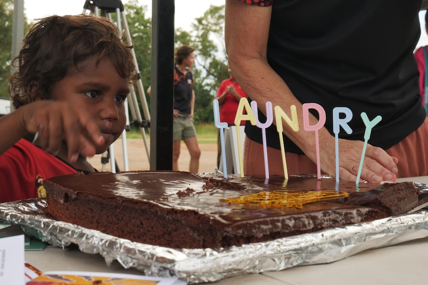 A kid looks on eagerly as a cake is cut. Candles in the shape of letters spell the word "laundry". 