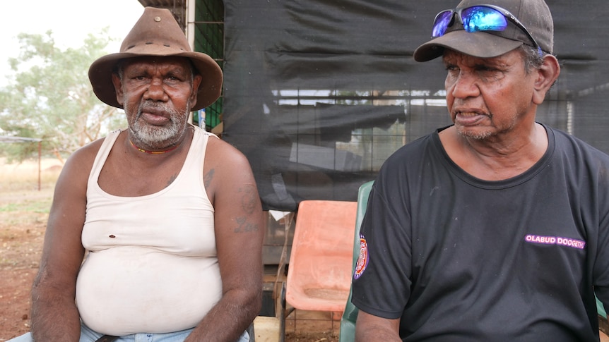 two indigenous men sit next to each other in front of a shed