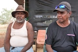two indigenous men sit next to each other in front of a shed