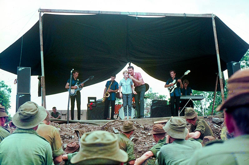 Little Pattie and Col Joye performing on stage to a crowd of uniformed soldiers.