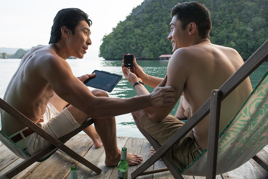 Colour still of actors Chris Pang and Henry Golding sitting on a wooden deck by the water in 2018 film Crazy Rich Asians.