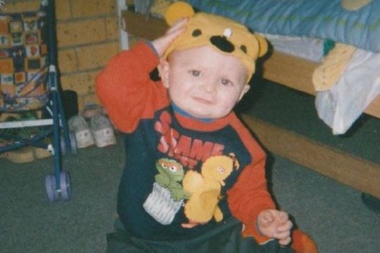 A toddler, wearing a Sesame Street top and a beanie sporting a nose, eyes and ears, smiles for the camera.