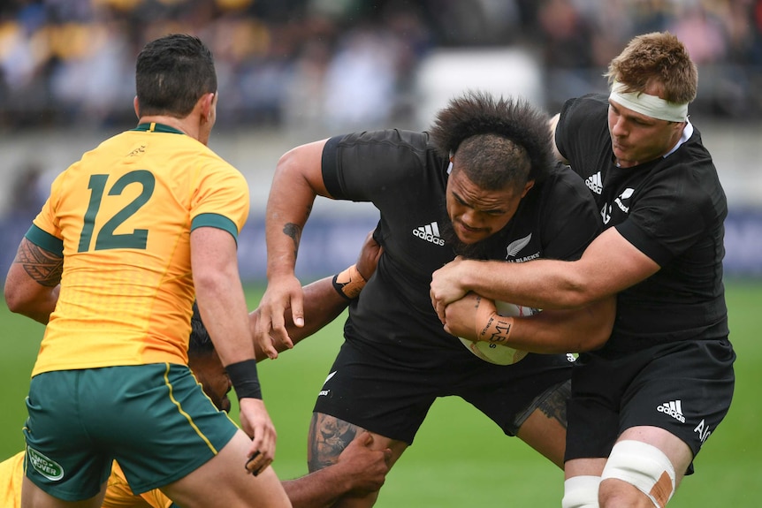 A New Zealand All Blacks players holds the ball while under pressure from the Australian defence.