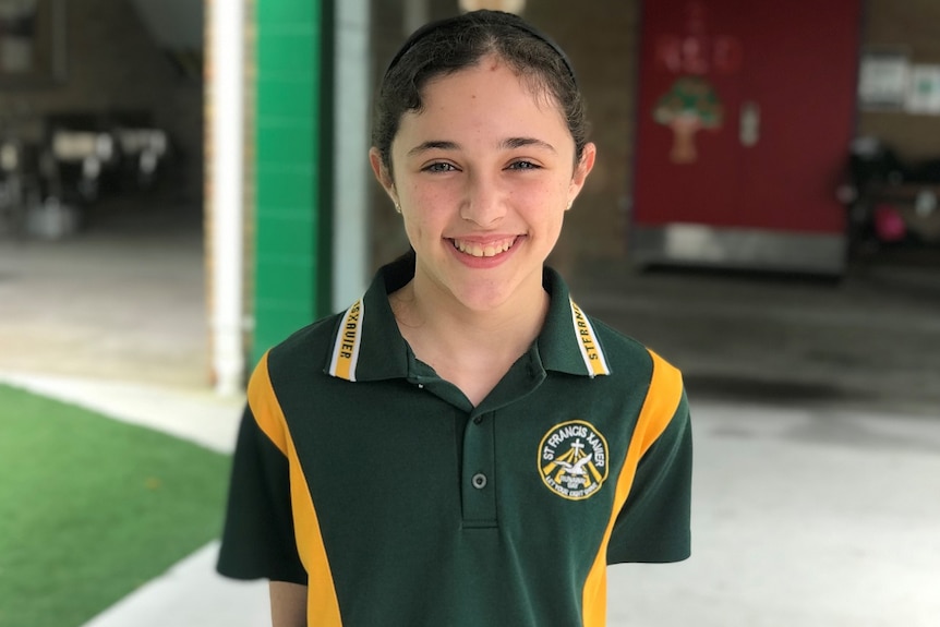 A young girl with tied back dark hair wearing a green and yellow t shirt smiling on a bright day