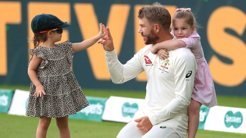 David Warner is hugged from behind by one daughters while the other, wearing his baggy green and sunglasses, touches his hand.
