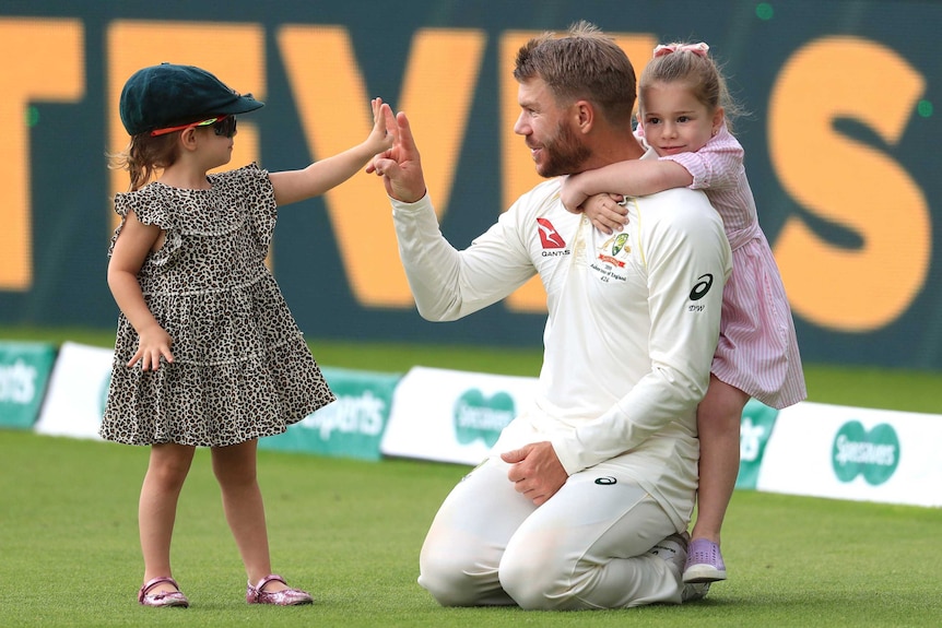 David Warner is hugged from behind by one daughters while the other, wearing his baggy green and sunglasses, touches his hand.